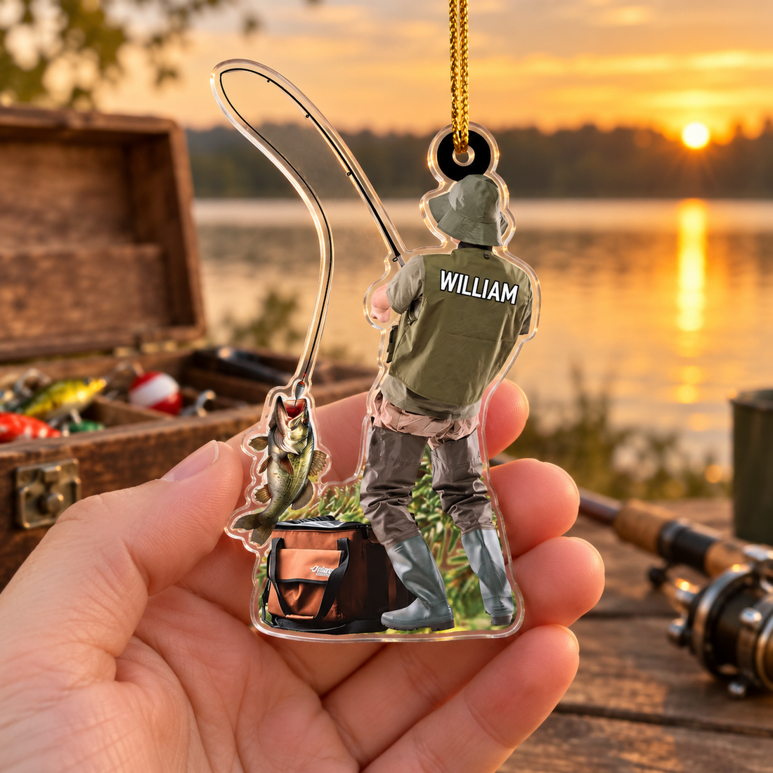 Hand holding a personalized fisherman William ornament in front of a tackle box and a lake at sunset