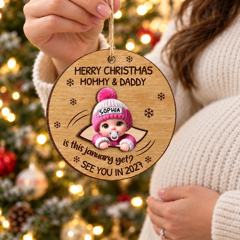 Expecting mom holding personalized baby peeking 2-layered wood ornament near Christmas tree