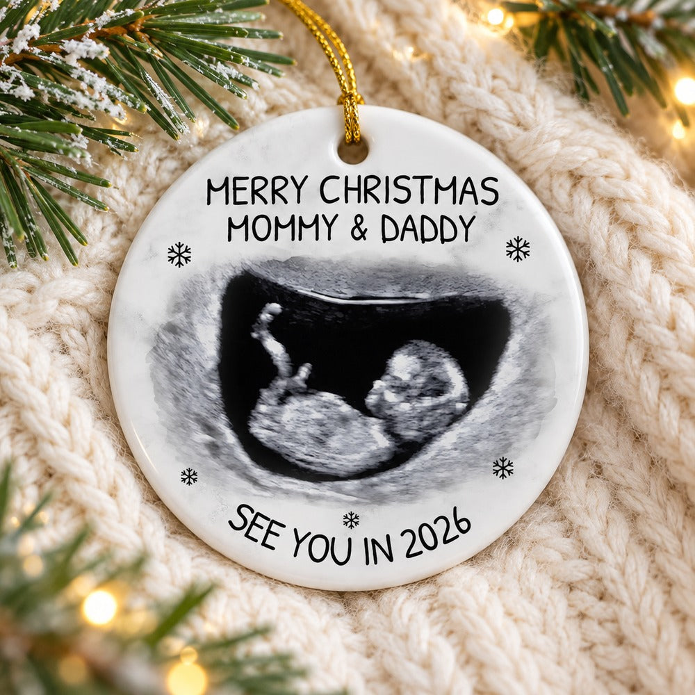 Personalized baby expecting 2026 ceramic ornament lying flat on a cozy cream knit blanket with pine sprigs