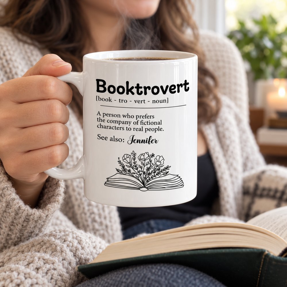 Book lover holding steaming personalized Booktrovert ceramic mug while reading a book in a cozy chair