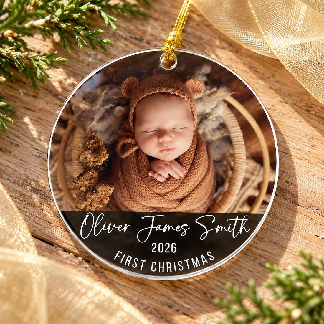 Custom photo baby first Christmas acrylic ornament flat on a wood surface with cedar greenery