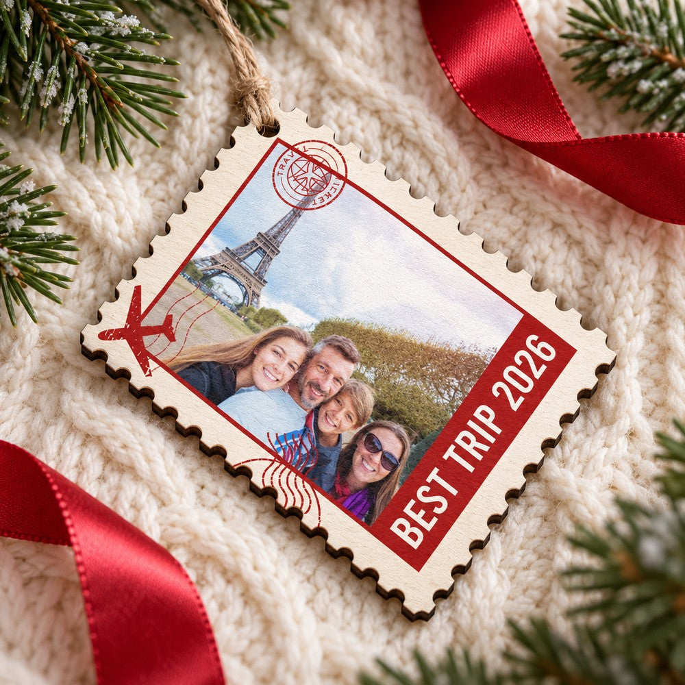 Personalized family travel stamp wood ornament lying flat on a cozy cream knit blanket with pine greenery