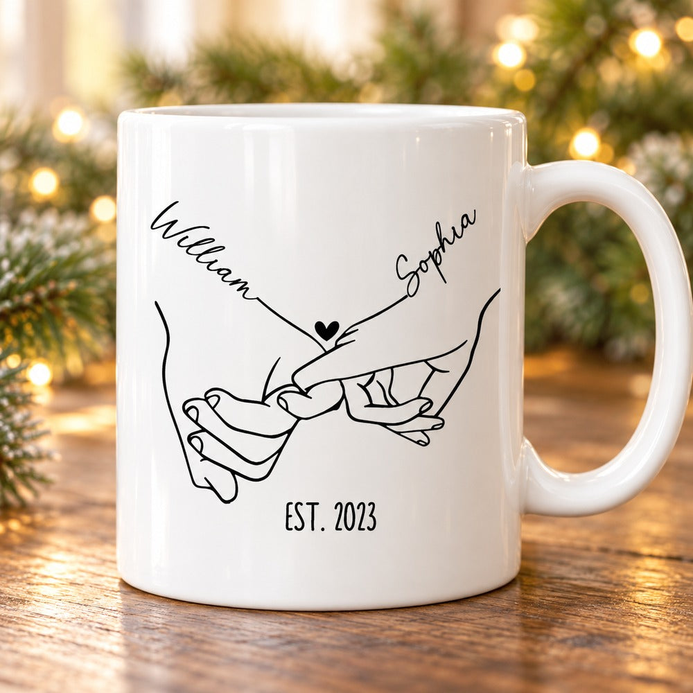 Personalized holding hands couple mug on a holiday table with pine greenery and fairy lights