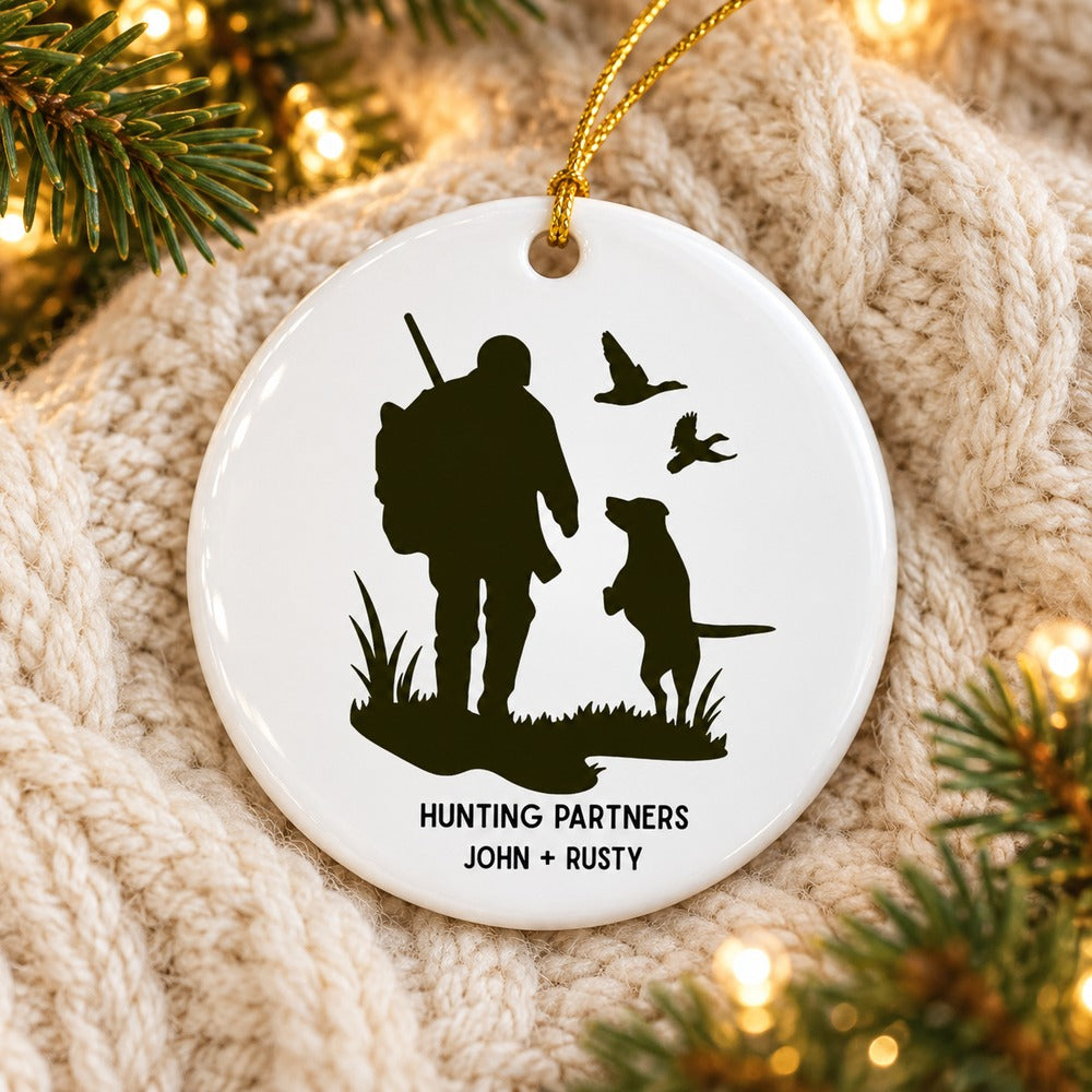 Custom hunter and dog ceramic ornament lying flat on a cream knit blanket with pine greenery