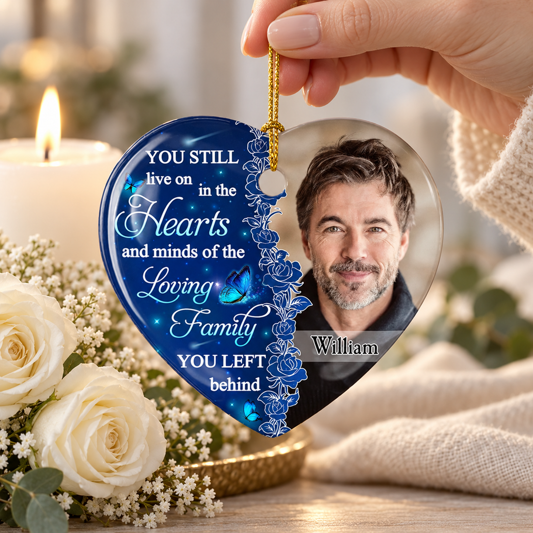 Hand holding a personalized heart memorial photo ornament beside a white candle and white roses