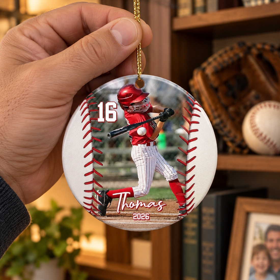 Hand holding a personalized baseball ceramic ornament in front of a bookshelf with a glove and ball