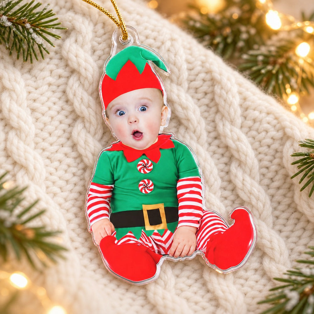 Custom photo baby elf acrylic ornament lying flat on a cozy cream knit throw with pine sprigs
