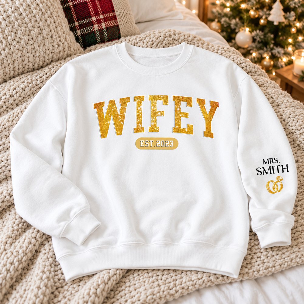 Personalized Wifey Est sweatshirt laid on a cozy bed with holiday tree background